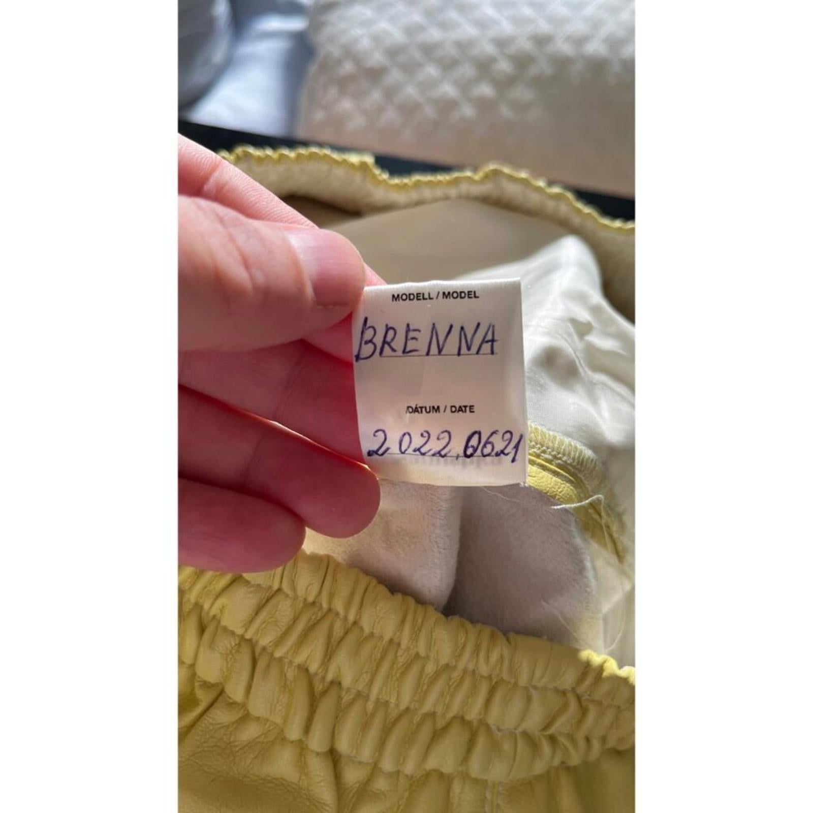Label showing model name 'BRENNA' from NANUSHKA yellow vegan leather elastic waist shorts.
