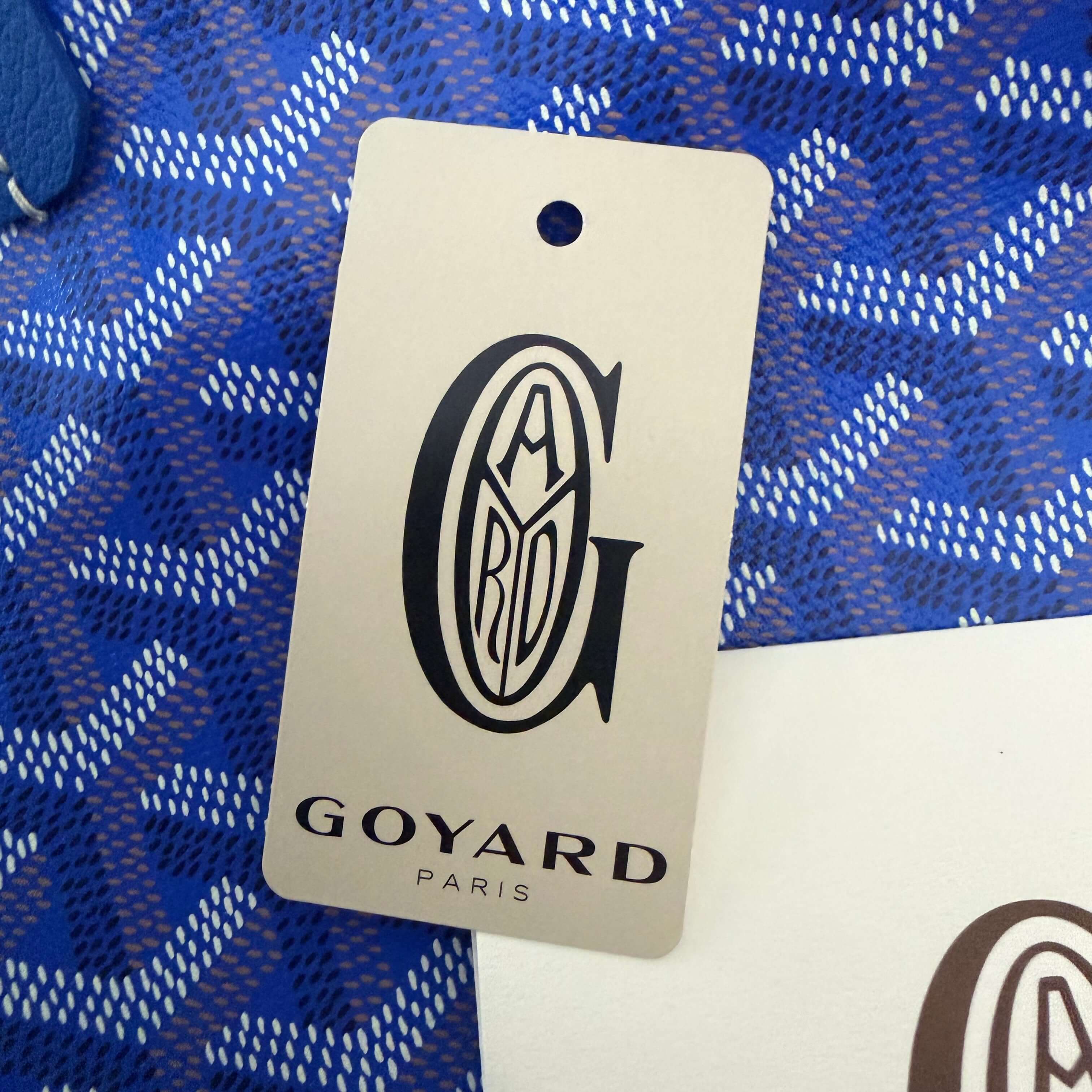 Goyard Artois MM Tote Bag- Goyardine tag featuring Goyard Paris logo on a blue background.